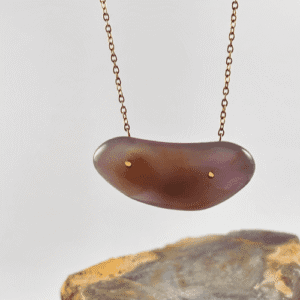 Collar con colgante de Calcedonia morada, lisa y curvada, con puntos dorados, colgando en el aire sobre una roca gris.