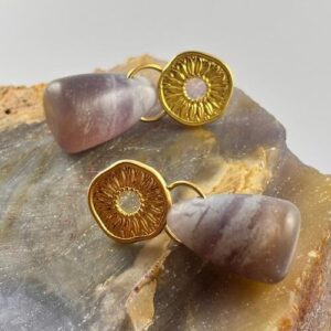Detalle en primer plano de aretes con piedra morada irregular colgando de un disco dorado.