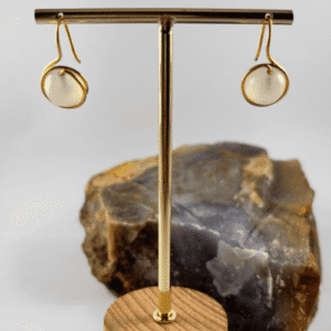 Par de aretes de calcedonia blanca redonda engarzada en alambre dorado, sobre ganchos dorados, exhibidos en un soporte de metal.