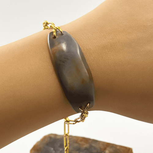 Pulsera de cadena dorada con una piedra plana ovalada de Calcedonia morada y beige, puesta en una muñeca.