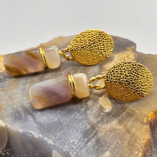 Pendientes de piedras cilíndricas jaspeadas moradas y beige, con cierre circular dorado , descansando sobre una calcedonia en bruto.