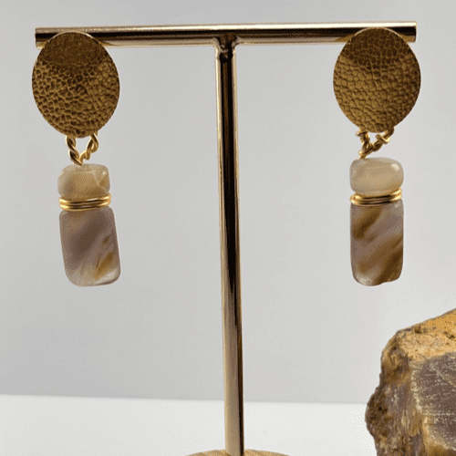 Aretes con poste circular dorado y piedra cilíndrica de amatista o ágata con bandas, en exhibidor de joyería.